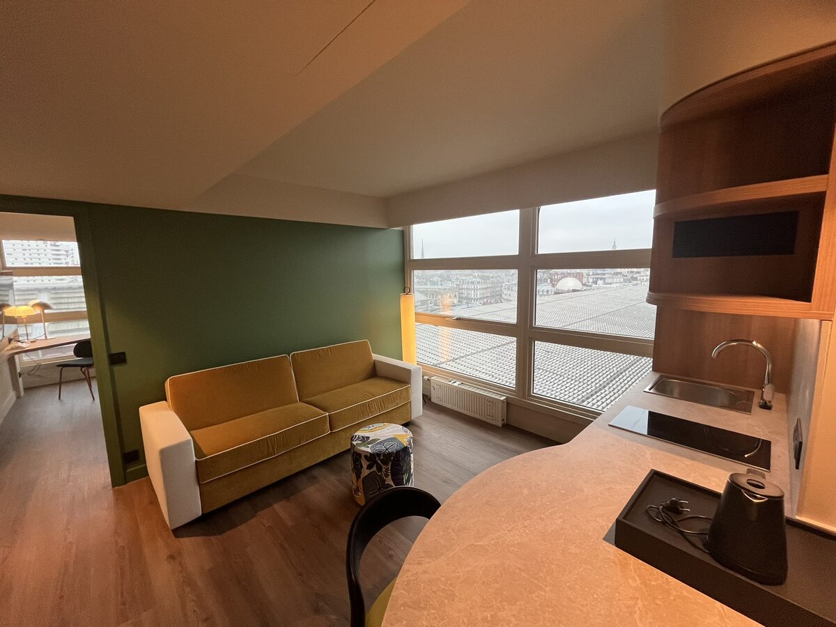 Espace salon avec canapé jaune, vue sur les toits et kitchenette intégrée